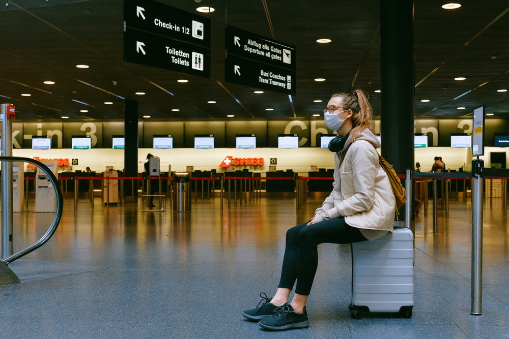 Image of a woman sitting on a suitcase while wearing a protective mask.